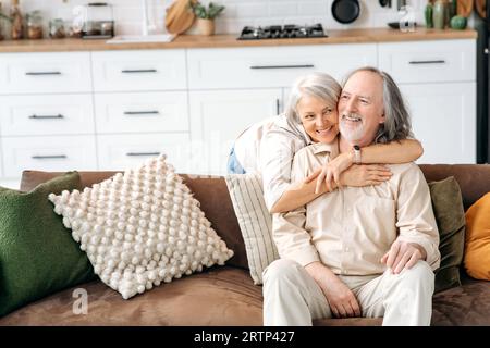 Photo of happy caucasian gray haired spouses of retirement age, husband and wife spend time together in living room, a woman hugs her beloved man sitting on a sofa, they smile, rejoice to be together Stock Photo