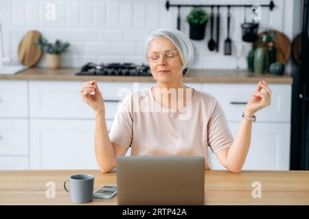 Meditation concept. Calm relaxed mature caucasian gray haired woman, sitting at home in the kitchen at the table with a laptop, relaxing while working online, meditating with her eyes closed Stock Photo