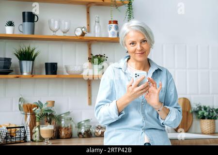 Positive stylish caucasian mature grey haired woman, stands at home in the kitchen, holds her smartphone in hands, uses it to communicate in social networks, read news, looks at the camera, smiles Stock Photo