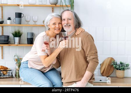Anniversary. Happy pensioners with wine. Middle aged caucasian spouses, sit at home in the kitchen, hold glasses of red wine in their hands, celebrate an important date, anniversary, smile at camera Stock Photo