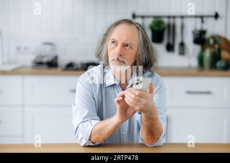 Thoughtful middle aged caucasian grey haired man with beard, sits at home in the kitchen at the table, using his smartphone, browsing social media, reading news, shopping online, pensively looks away Stock Photo