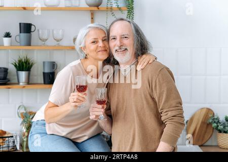 Photo of happy pensioners with wine. Middle aged caucasian spouses, sit at home in the kitchen, hold glasses of red wine in their hands, celebrate an important date, anniversary, smile at camera Stock Photo