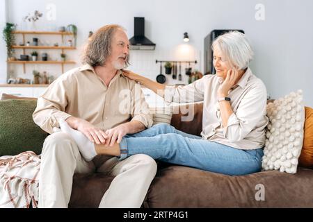Caucasian gray haired pensioners, mature husband and wife, spend time together at home on the sofa in the living room, man massaging his beloved wife feet, they resting and relaxing together Stock Photo