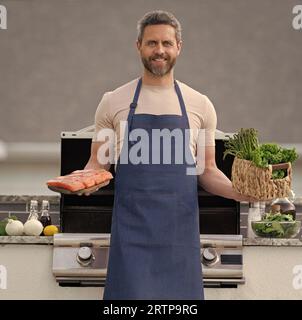 photo of glad barbecue man with salmon fish. barbecue man with salmon. barbecue man with salmon outdoor. barbecue man with salmon in apron. Stock Photo