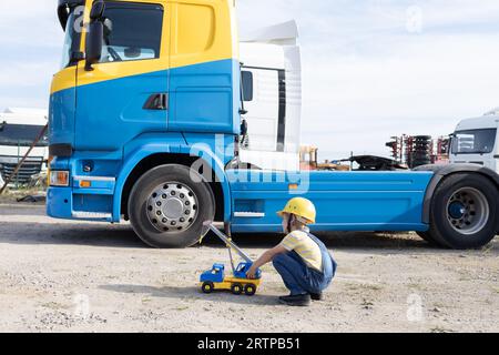 boy in construction helmet with toy car plays with interest while sitting on construction site in front of dump truck cabin. Game in the profession. B Stock Photo