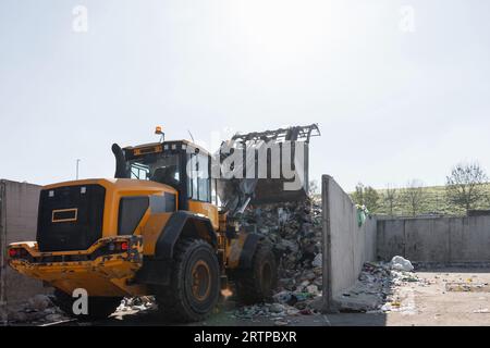 Yellow skid steer loader moving wooden waste material, shaking out a scrap grapple on the garbage heap in the materials recovery facility Stock Photo