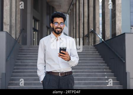 Successful young Hindu man walking outside office building, engineer software developer programmer smiling and happy using test app on phone, happy satisfied with result. Stock Photo