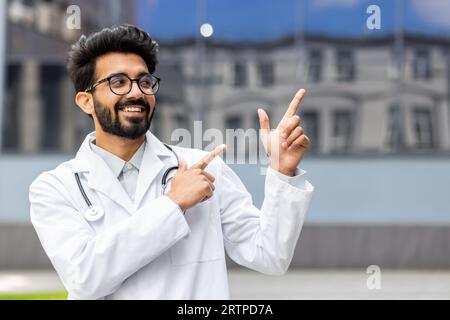 A young Hindu doctor from outside the clinic smiles and looks away, the man points his fingers in the direction, smiles with satisfaction and the doctor with a medical coat and a stethoscope. Stock Photo
