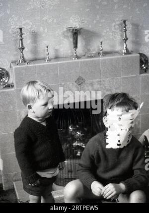 1960s, historical, what's that funny thing on your face....a little boy looking at this brother with his home made paper mask on his face, as they sit infront of a tiled fireplace of the era, England, UK. Stock Photo