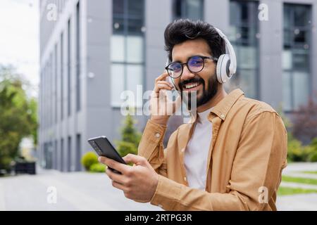 Portrait of a young indian programmer outside an office building on a work break, a man in headphones listens to music, smiles and looks at the camera, a businessman uses an application on phone. Stock Photo
