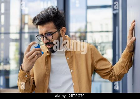 Young man coughing with asthma, businessman outside office building using inhaler. Stock Photo