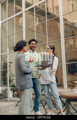 Cheerful coworkers discuss the project while use laptop standing on cafe terrace background Stock Photo