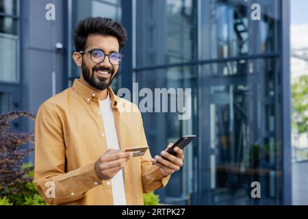 Portrait of young man outside office building, hispanic smiling and looking at camera, holding phone and bank credit card, online shopping and booking services remotely. Stock Photo