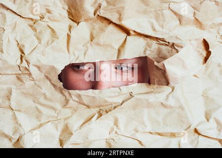 female eyes looks  hole in a crumpled paper background. Woman peeking out of hole in paper Stock Photo