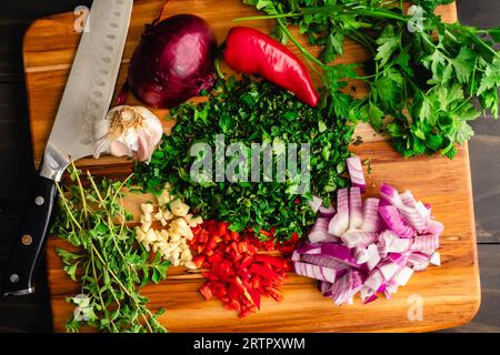 Prepping Chimichurri Ingredients on a Wooden Cutting Board: Chopped parsley, mint, and oregano with red onion, garlic, and red chili pepper Stock Photo