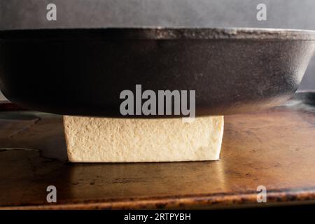 Pressing a Block of Tofu with a Cast Iron Skillet: Pressing the water out of a block of bean curd using a heavy pan Stock Photo