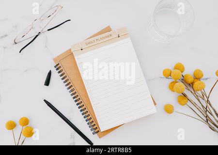 Flat lay of white notebook with copy space for text, glasses and yellow flowers on white marble background. Top view. Minimal home office desk. Work o Stock Photo