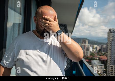 Man hiding from sun covering face with hand standing on balcony in city in extra hot weather. Stock Photo