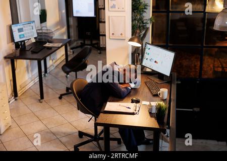 Executive manager doing overtime work in startup office, falling asleep desk trying to finish marketing strategy for company project. Businessman working late at night, sleeping on table at workspace Stock Photo