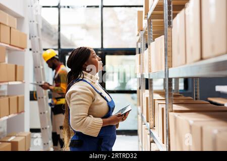 Logistics and distribution manager inspecting merchandise storage in warehouse. African american storehouse employee standing near parcels shelf, holding tablet and looking at carton Stock Photo