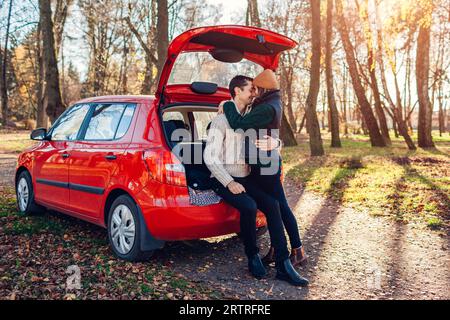 Happy loving couple relaxing in car trunk hugging in autumn forest. Man and woman travel by auto wearing warm clothes. Young people enjoy fall trip. S Stock Photo