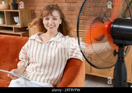 Young woman with tablet computer and blowing electric fan sitting on sofa at home Stock Photo