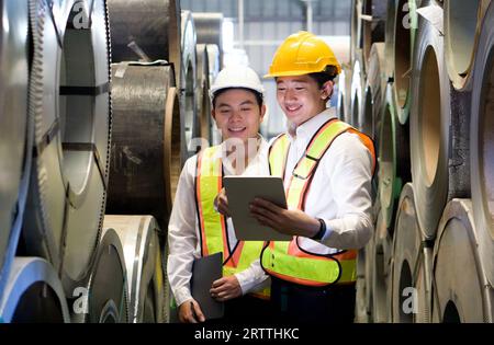 Two dedicated workers, adorned with protective hard hats, stand confidently in the industrial arena, surrounded by massive steel coils. Stock Photo