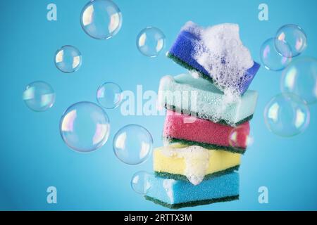Stack of kitchen multi coloured sponges with detergent foam and soap bubbles on blue background. Creative dish washing sponges. Stock Photo