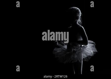 A young female dancer wearing a white tutu stands in a dark studio, her arms raised in a graceful pose Stock Photo