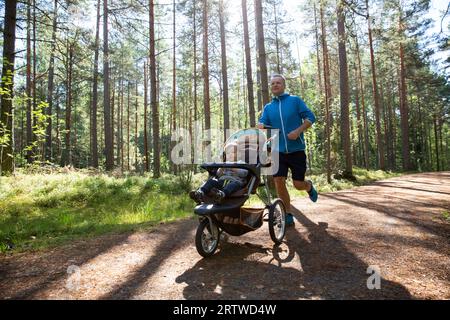 Athletic man in sportswear running with baby stroller at sunny morning in city park during fatherhood time. Stock Photo
