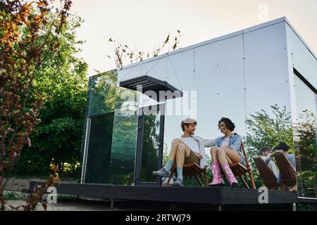 happy multiethnic couple sitting on chairs and looking at each other near modern glass house Stock Photo