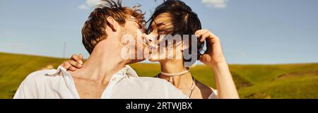 rustic wedding, young interracial newlyweds kissing on wind under blue sky in green field, banner Stock Photo