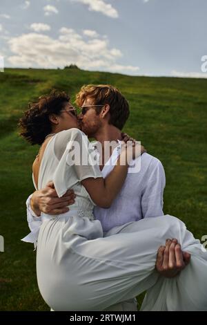 happy newlyweds,  groom lifting and kissing asian bride in white dress in green field, countryside Stock Photo