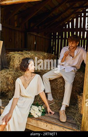 happy asian bride in white dress holding bouquet and sitting on hay near groom in barn, rural Stock Photo