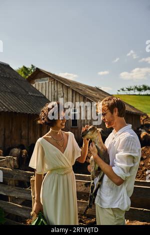 happy multiethnic newlyweds in wedding gown and sunglasses cuddling cute baby goat, countryside Stock Photo