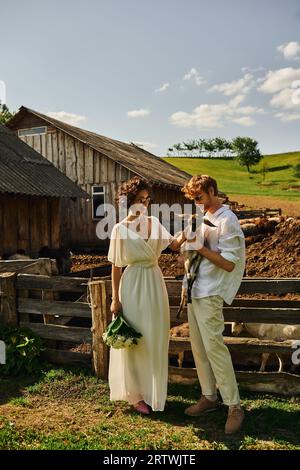 happy multiethnic couple in wedding gown and sunglasses cuddling cute baby goat, countryside Stock Photo