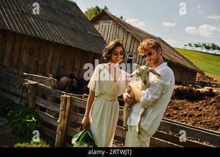 smiling multiethnic couple in wedding gown and sunglasses cuddling cute baby goat, countryside Stock Photo