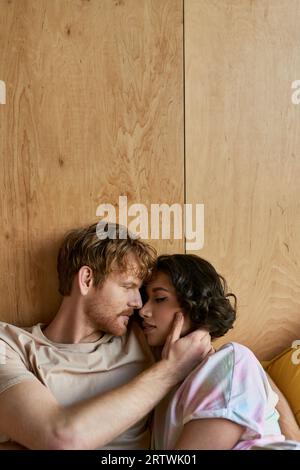 interracial couple having tender moment, redhead man touching cheek of asian woman and lying on bed Stock Photo
