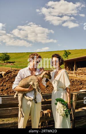 newlyweds in countryside, happy man holding goat near asian bride in white dress, rustic wedding Stock Photo