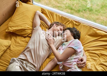 top view of happy man hugging asian girlfriend and lying together on yellow bedding at home, couple Stock Photo