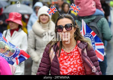 A woman with Union Jack flags, walks down Piccadilly in the rain, part of the large crowd heading to watch King Charles III’s coronation. Piccadilly, Stock Photo