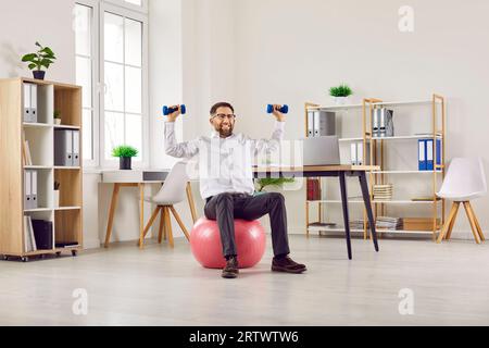 Funny young business man doing sport exercises on fit ball with dumbbells in office. Stock Photo