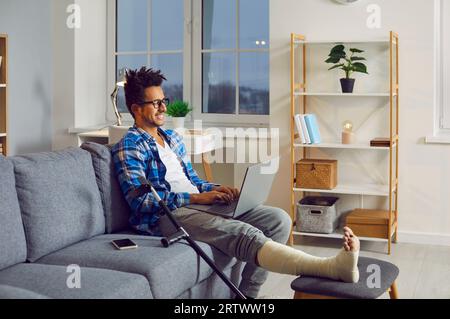 Young man with broken leg in plaster cast sitting on couch with laptop Stock Photo