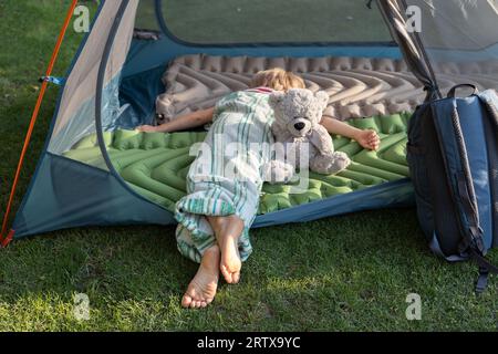 tired barefoot child sleeps or rests lying on his stomach in a tent with his favorite toy teddy bear. Healthy lifestyle, little tourist, family activi Stock Photo