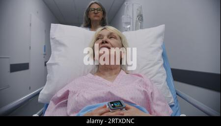 Mature female doctor with young nurse brings senior patient to medical center. Old woman lies on gurney with drip and talks with medics about surgery. Medical staff at work in modern hospital. Stock Photo