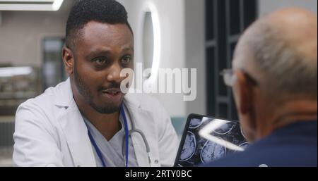 African American doctor sits in clinic cafe with elderly patient. Health care specialist talks to senior man, shows MRI or CT brain scan image using digital tablet. Hospital or medical center canteen. Stock Photo