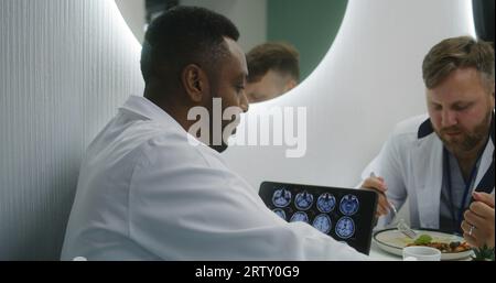 African American doctor sits in clinic cafe with colleagues. Healthcare specialists talk during lunch. Digital tablet with MRI brain scan image. Medical staff in hospital or medical center canteen. Stock Photo