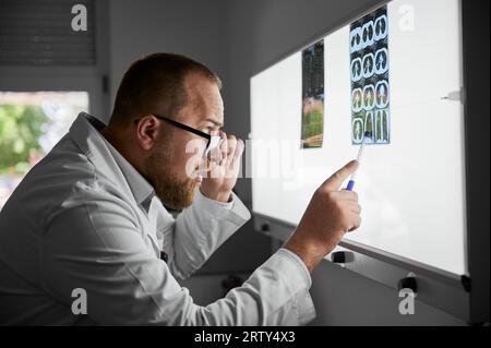 Side view of serious doctor studying MRI results. Male specialist working in hospital, carrying out description of radiographs of patient, pointing by pen. Concept of medicine and hospital. Stock Photo