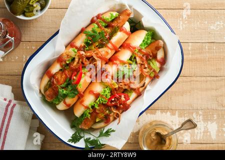 Delicious grilled hotdog with pickled cucumbers, chili peppers, caramelized onions and ketchup, mustard in craft paper on old wooden background. Homem Stock Photo