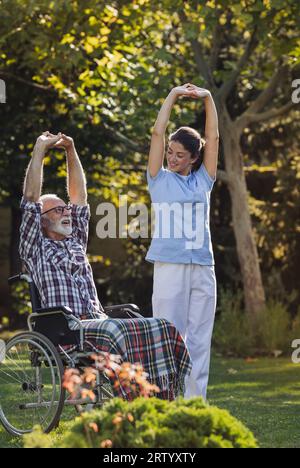 Senior man with disability in wheelchair stretching arms and doing workout with young woman caregiver and physiotherapist in park Stock Photo